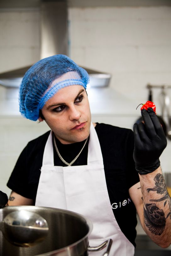 Kier Kemp, who started the Condimaniacs brand in Kent, is seen in front of a large cooking pan in an industrial kitchen setting. He is dressed in catering wear and is inspecting ingredients which include a chilli pepper