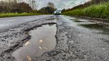 A picture of a pothole in a road - in the distance there is a car driving forwards.