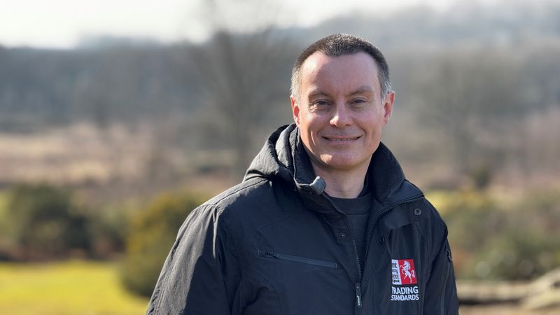 Kent Trading Standards' Kieron Watts in a black jacket uniform with Kent County Council logo standing in the Kent countryside