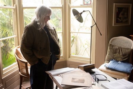 Deirdre Kashdan standing in a dark room in front of a bay window looking down at an office desk covered in artwork and books 