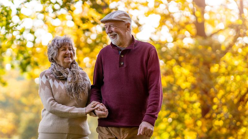 Older woman with a man, wearing a flat cap, walking past a tree with glowing yellow and orange autumnal leaves 