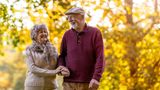 Older woman with a man, wearing a flat cap, walking past a tree with glowing yellow and orange autumnal leaves 