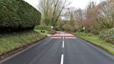 A newly resurfaced road surrounded by grass verges and trees. There are red traffic calming measures painted onto the road next to national speed limit signs 