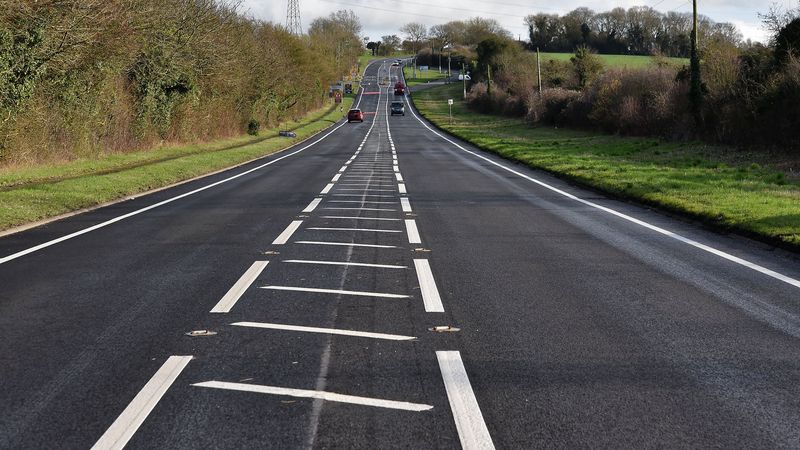 A long, freshly resurfaced two‑lane road with bright white road markings stretching into the distance through a rural area. Several cars are travelling in both directions, and the road is bordered by grass, hedges, and trees under an overcast sky