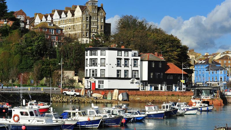 Harbour scene with several small boats moored in calm water, with a row of colourful waterfront buildings and larger houses on a hillside in the background under a bright, partly cloudy sky