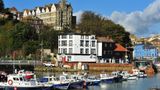 Harbour scene with several small boats moored in calm water, with a row of colourful waterfront buildings and larger houses on a hillside in the background under a bright, partly cloudy sky