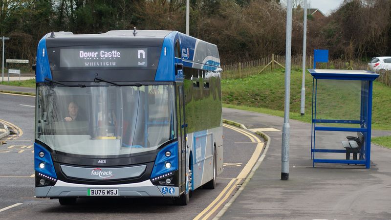 A modern dark blue and silver Fastrack bus marked for Dover Castle, driving along a curved road near a small shelter and surrounded by grassy banks and fencing.