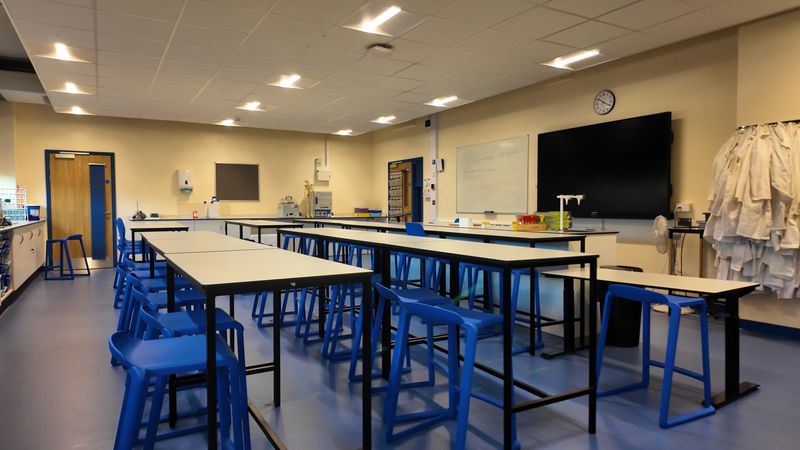 An empty science classroom with long white laboratory tables, blue stools, and equipment benches along the walls. The room includes a large digital screen, a whiteboard, storage cabinets, and several lab coats hanging on hooks. Overhead lights illuminate the space.