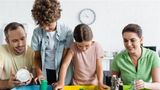 Four people sorting household waste into colour‑coded recycling bins, placing items such as cans, bottles and packaging into separate containers in an indoor setting.