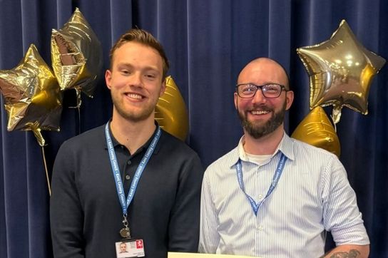 Two people standing side by side in front of a dark blue curtain, wearing lanyards and smiling at the camera. Several gold, star-shaped balloons are positioned behind them