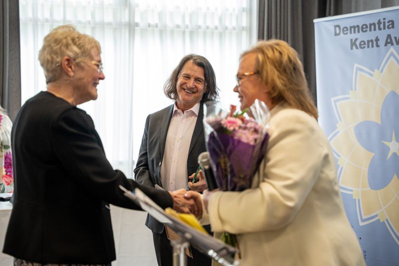A person receiving an award and flowers in front of a Dementia Friendly Kent Awards branded banner