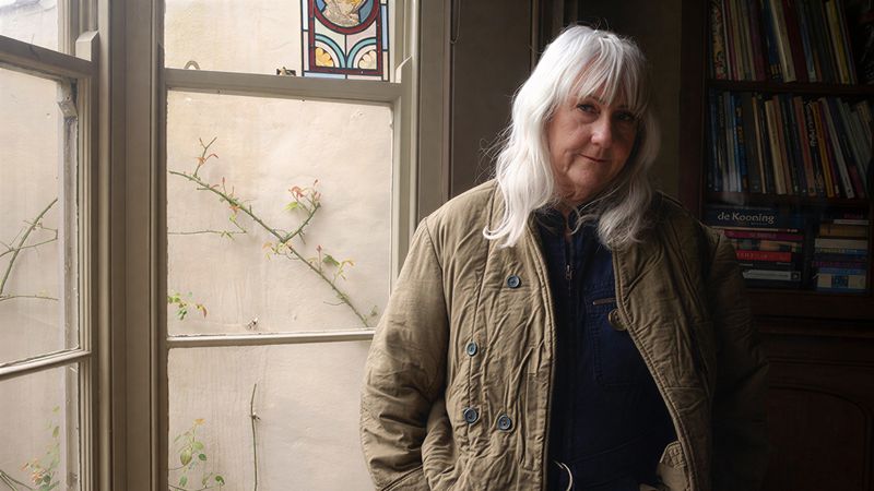 A woman with grey hair stands with her hands in her pockets looking toward the camera.
