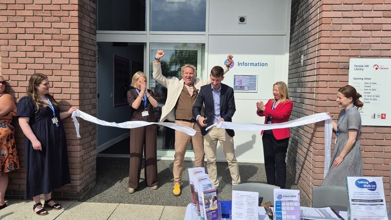 A group of people stand outside a brick building entrance during a ribbon-cutting ceremony. One person cuts a long white ribbon with scissors while others stand on either side holding the ribbon and clapping. Behind them, a sign on the building reads &ldquo;Information,&rdquo; and a display table with leaflets is set up in the foreground.