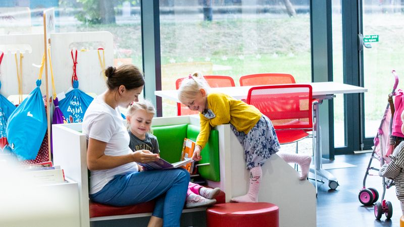 An adult shares a book with two children in a library reading area, encouraging reading and storytelling together.
