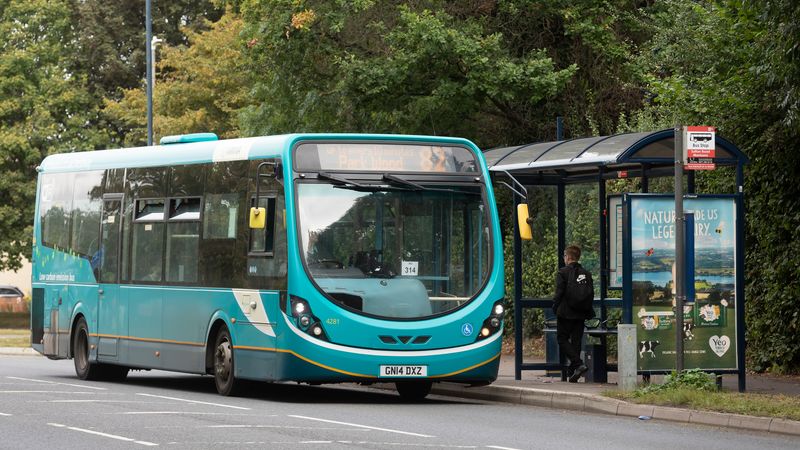 A turquoise Arriva bus on route 82 to Park Wood is stopped at a bus shelter on Sutton Road. A person stands near the shelter, which displays a large advertisement, while trees and greenery line the roadside in the background.