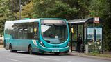 A turquoise Arriva bus on route 82 to Park Wood is stopped at a bus shelter on Sutton Road. A person stands near the shelter, which displays a large advertisement, while trees and greenery line the roadside in the background.