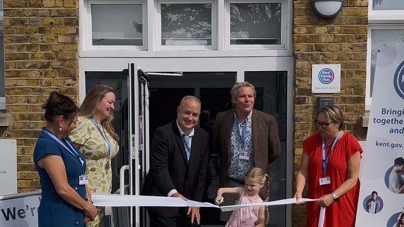 People stand outside the entrance of Queenborough Library and Family Hub during a ribbon-cutting ceremony. A young child in pink clothing cuts a white ribbon with scissors while several adults hold the ribbon and stand nearby. The group is positioned in front of the building's glass entrance doors. Signs for the library reopening and Kent Family Hub are visible on either side of the doorway.
