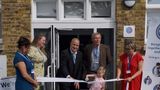 People stand outside the entrance of Queenborough Library and Family Hub during a ribbon-cutting ceremony. A young child in pink clothing cuts a white ribbon with scissors while several adults hold the ribbon and stand nearby. The group is positioned in front of the building's glass entrance doors. Signs for the library reopening and Kent Family Hub are visible on either side of the doorway.