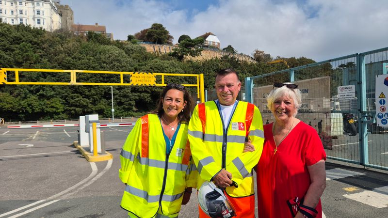 Three people stand at the entrance to the Road of Remembrance work site, with the cliff stabilisation area visible in the background. Two people are wearing high-visibility safety jackets, and fencing and site infrastructure can be seen around the entrance.