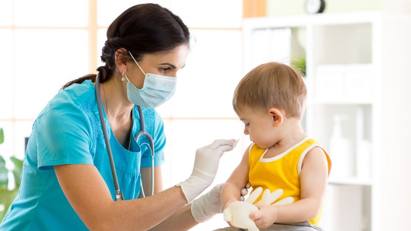Child in bright yellow top looks at his arm as a nurse prepares to give him his vaccination