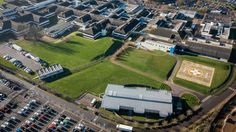 A drone image of a hospital building. There is a big H on the ground. Lots of houses are in the top of the photo.