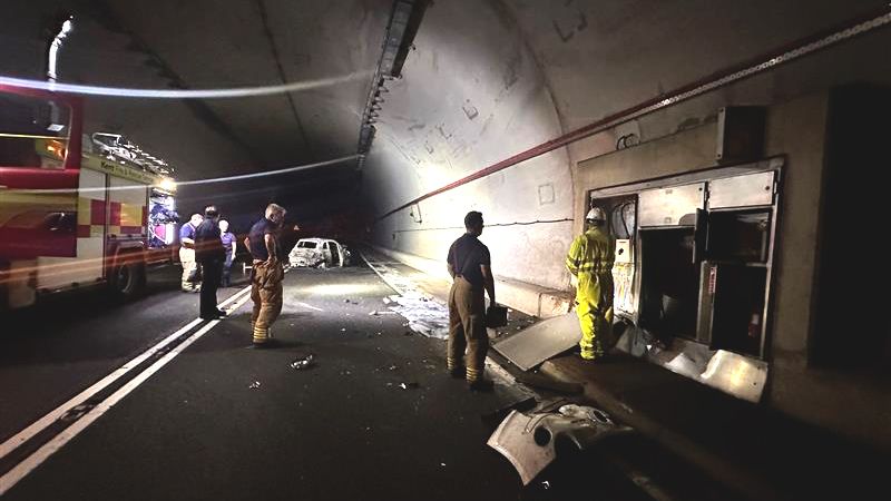 The photograph is taken inside a dark tunnel. Figures are looking at electricals and on the left is a red fire engine