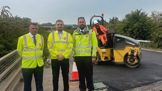 Three men look towards the camera in hi-vis. Behind them is a roller on the road. 