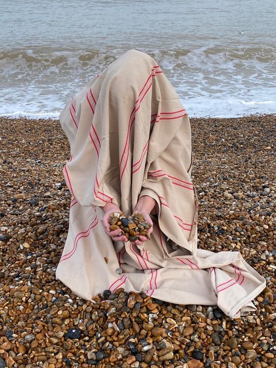 A woman covered in a bedsheet crouched on a pebble beach next to the sea. She is holding out pebbles in her hand which are sticking out from under the sheet