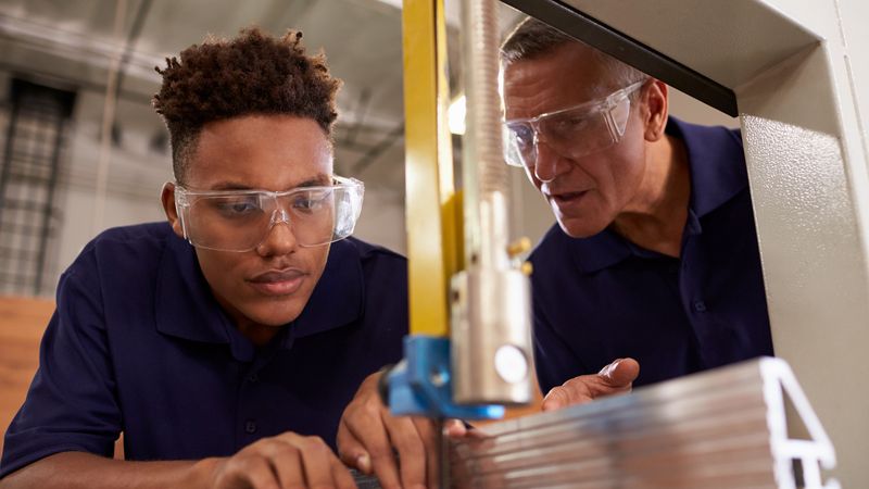 A young apprentice wearing safety goggles and overalls being taught how to use a machine by an older mentor. They are leaving over a workbench concentrating. 