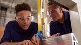 A young apprentice wearing safety goggles and overalls being taught how to use a machine by an older mentor. They are leaving over a workbench concentrating. 