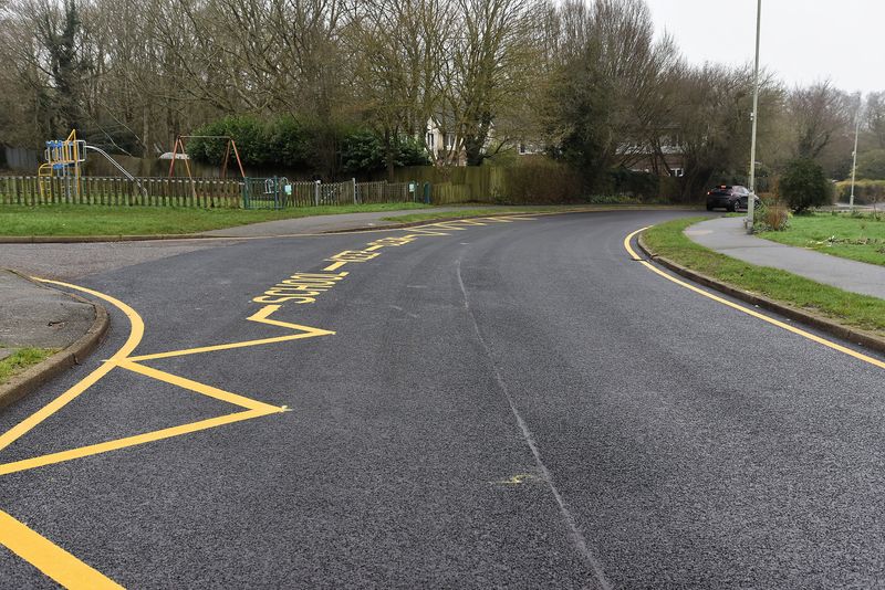 A curved residential road with new bright yellow road markings, including a zigzag &lsquo;School Keep Clear&rsquo; zone. A children&rsquo;s playground with swings sits behind a low fence to the left, with houses and trees in the background.