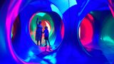 The interior of an immersive, inflatable art structure illuminated with vivid blue, pink, green, and red light. Two people stand inside the curved, tunnel‑like space, surrounded by circular openings that connect to other glowing chambers.