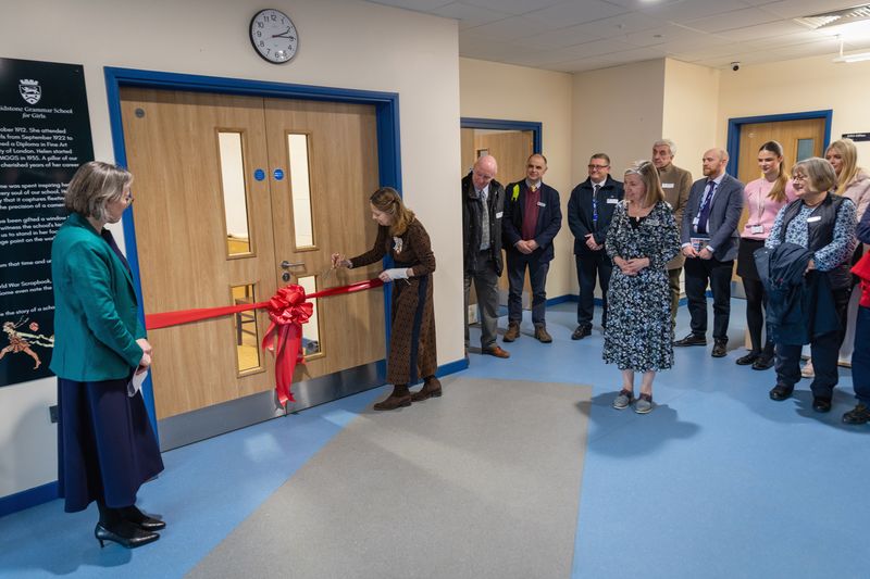 A group of people stand in a hallway watching a ribbon‑cutting ceremony in front of a pair of wooden double doors. One person cuts a large red ribbon that is tied in a bow across the doors, while others stand nearby observing. The hallway has blue flooring, cream walls, and a clock above the doors.