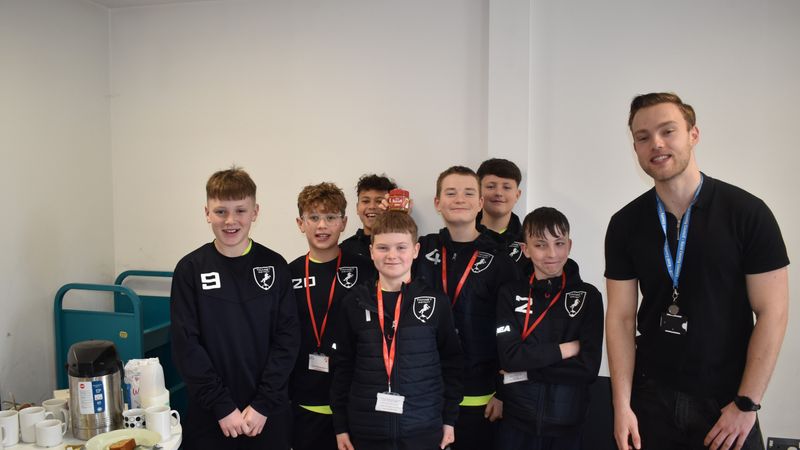 A group of young football players wearing matching dark sports kits and lanyards standing together indoors, with an adult wearing a lanyard standing to one side. A table with cups, a hot drinks flask, and plates of food is visible nearby, suggesting a community or team event in a library or indoor venue.