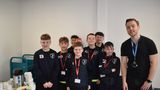 A group of young football players wearing matching dark sports kits and lanyards standing together indoors, with an adult wearing a lanyard standing to one side. A table with cups, a hot drinks flask, and plates of food is visible nearby, suggesting a community or team event in a library or indoor venue.