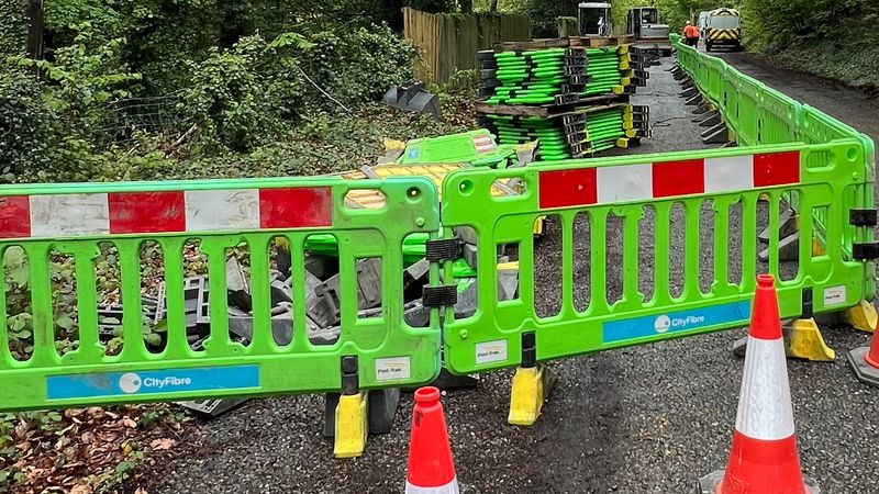 Roadworks on a narrow, tree-lined lane, with bright green safety barriers and orange cones blocking the foreground. Stacks of green panels and tools sit on the road, while small construction vehicles and two workers are visible further along the fenced-off area.