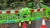 Roadworks on a narrow, tree-lined lane, with bright green safety barriers and orange cones blocking the foreground. Stacks of green panels and tools sit on the road, while small construction vehicles and two workers are visible further along the fenced-off area.