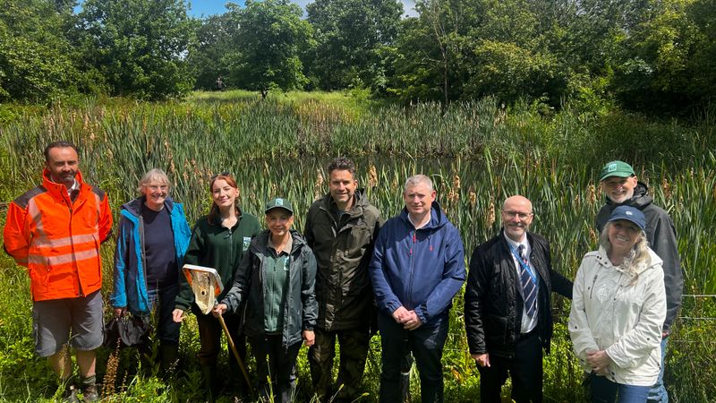 Group of people standing beside a reed-filled pond and wetland habitat during a site visit, with trees and vegetation in the background.