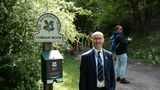 Person standing beside a National Trust sign for Cobham Wood on a tree-lined path, while several people walk along the woodland trail in the background.