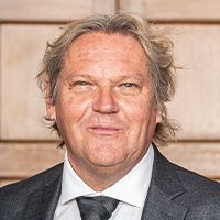 Paul Webb smiling for his photo in a dark blue suit, white shirt and dark blue tie infront of a bricked building