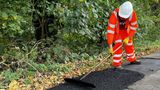 A highways worker wearing high-visibility orange safety clothing, gloves and a white hard hat uses a long-handled rake to spread fresh asphalt during a road repair. The worker is standing on a rural roadside next to dense green trees and fallen leaves.