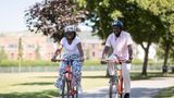 Two people wearing cycle helmets ride orange bicycles side by side along a paved park path. Trees provide shade over the path, with grass and blurred buildings visible in the background on a sunny day.