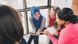 A group of young people are sitting and smiling, discussing something on a tablet one of them is holding