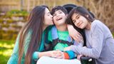 Two teenage sisters sit either side of their brother in a wheelchair and one kisses him on the cheek