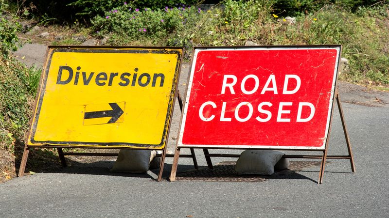 Two road signs on a road. The one on the left is a red rectangle with the words 'road closed' in the centre. The other sign is a yellow rectangle with the words 'diversion'.