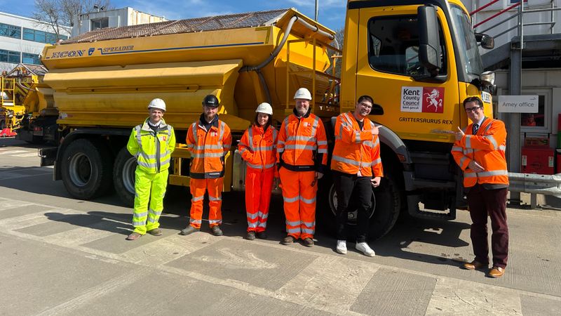 A group of people in hi-vis stand next to a big yellow gritter lorry. Two of them are pointing at a sign on the lorry which says 'Grittersaurus Rex'