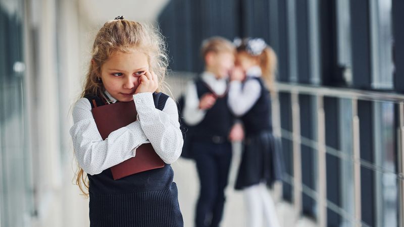 Young girl in school uniform looking sad while two children in the background are whispering