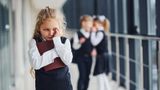 Young girl in school uniform looking sad while two children in the background are whispering