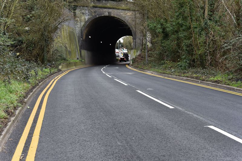 A recently resurfaced road with double yellow lines running towards a large brick tunnel beneath a railway bridge. Vegetation and trees surround the roadside, and a few vehicles are visible emerging from the tunnel