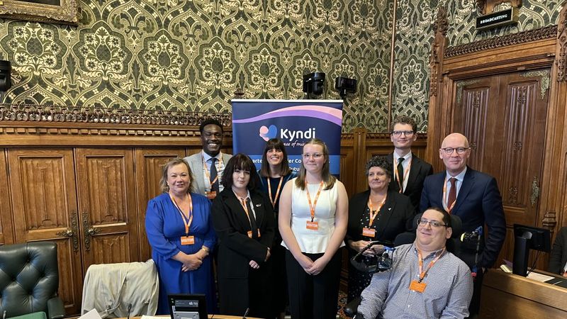 A group of nine people in a wood-paneled room with ornate patterned walls, seated desks in the foreground, and a &ldquo;Kyndi&rdquo; banner behind them.